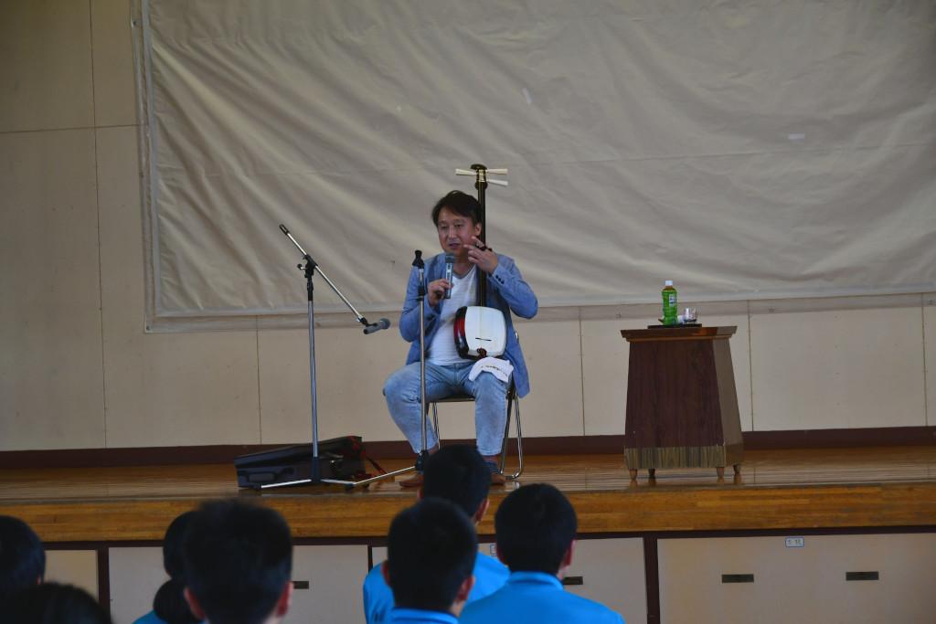 舞台上で椅子に座り三味線を持って話をしている松田さんの写真