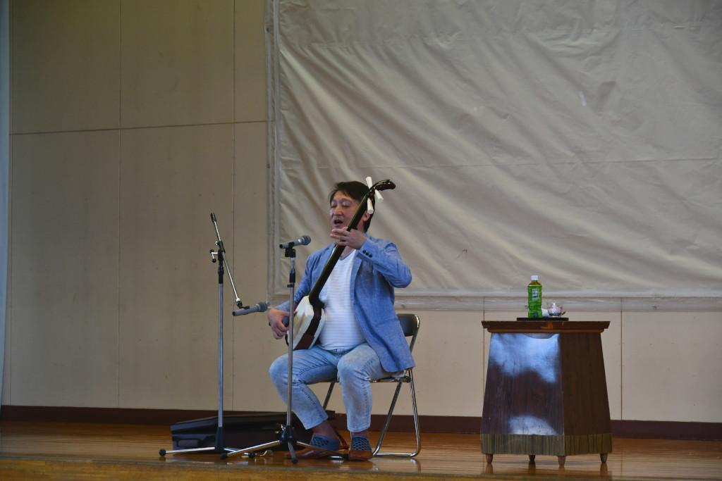 舞台上で椅子に座って三味線を弾きながら歌っている松田さんの写真