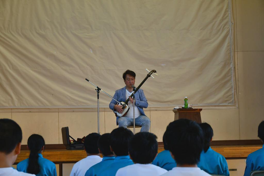 手前に演奏を聴いている生徒たちの後ろ姿と、舞台上で三味線を弾いている松田さんの写真