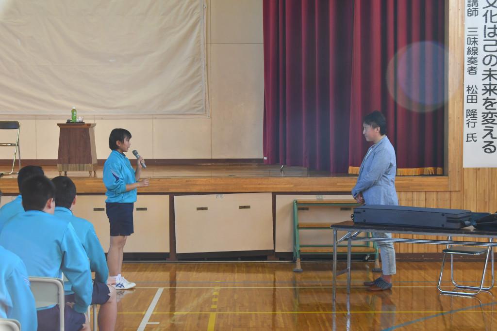 前に立ってあいさつをしている女子生徒と、向かい合う位置に立って話を聞いている松田さんの写真