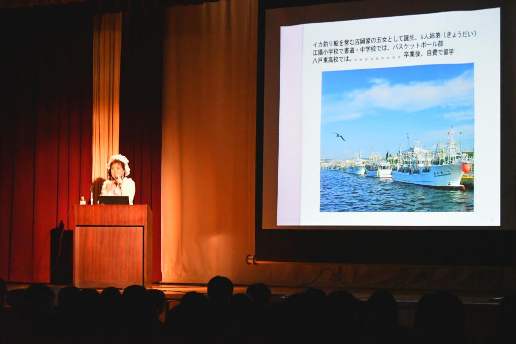 舞台の上、スクリーン横の演台の前で話をしている吉岡さんの写真