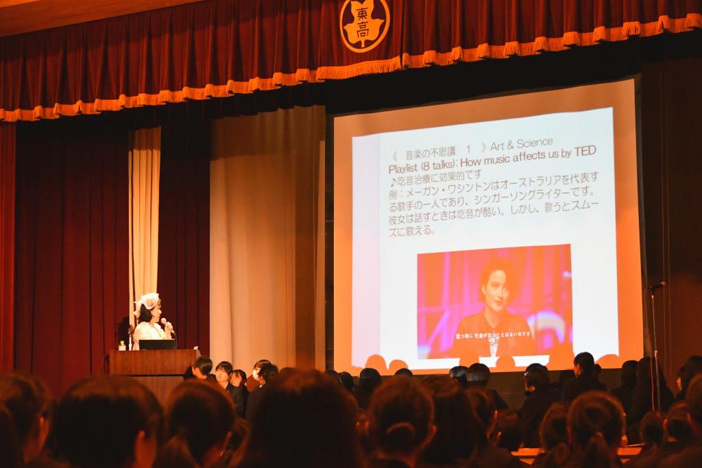 人物の写真と説明文の写ったスクリーンを見ながら演台の前に立って話をしている吉岡さんを客席後方から撮影した写真