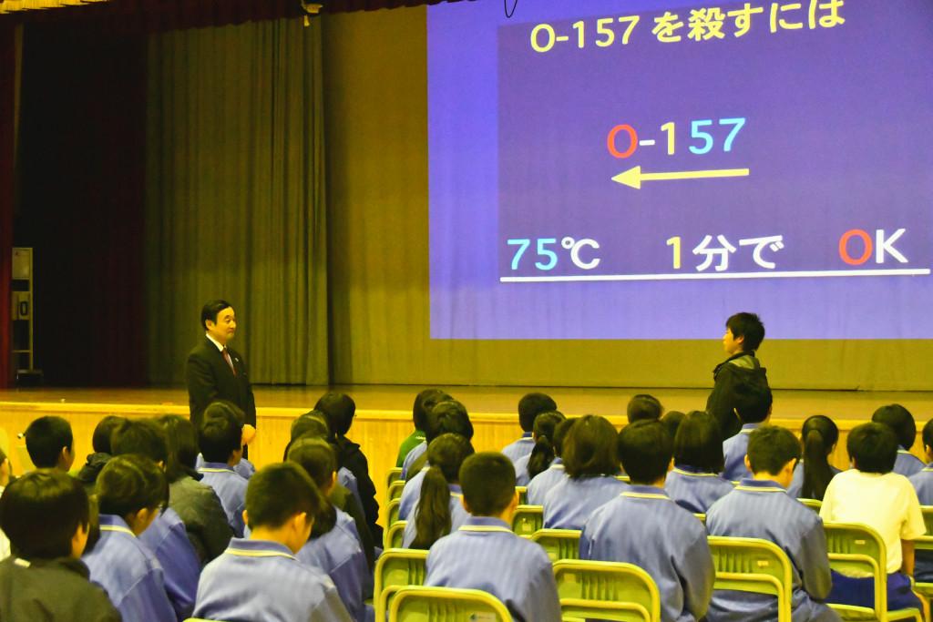 着席している生徒たちの前で代表であいさつをしている男子生徒と、向かい合う位置に立って話を聞いている野田先生の写真