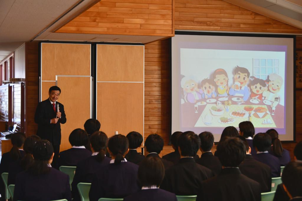 手前に着席して話を聞いている生徒の後姿、食事をしている家族のイラストが写っているスクリーンの横に立って話をしている野田先生の写真