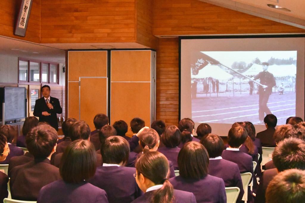 手前に着席して話を聞いている生徒の後姿、白黒の人物写真の写ったスクリーンの横に立って話をしている野田先生の写真