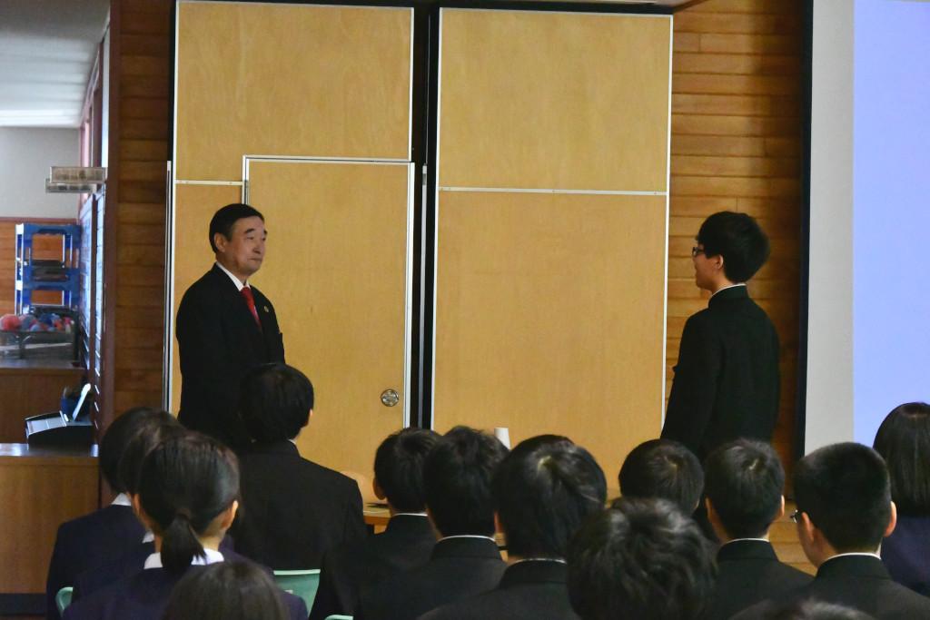 代表であいさつをしている男子生徒と、話を聞いている野田先生の写真