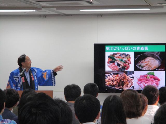 椅子に座る参加者たちを前に、モニターに映し出される青森の名物料理を紹介する男性の写真