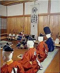 室内で、奥に鎧兜を身に着けた人が座り、手前に4人の人が昔の武士の衣装を着て座っている後姿の写真