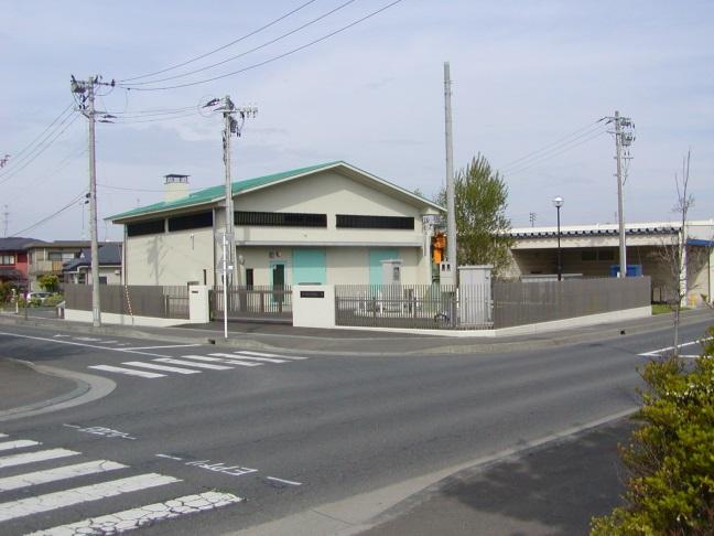 T字路の角にフェンスで囲まれエメラルドグリーンの切妻屋根とドアがある新井田西汚水中継ポンプ場の写真