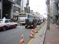 車道右側の歩道に赤と白の三角コーンが1列に並び、車道にバスやタクシーが走っている様子の写真