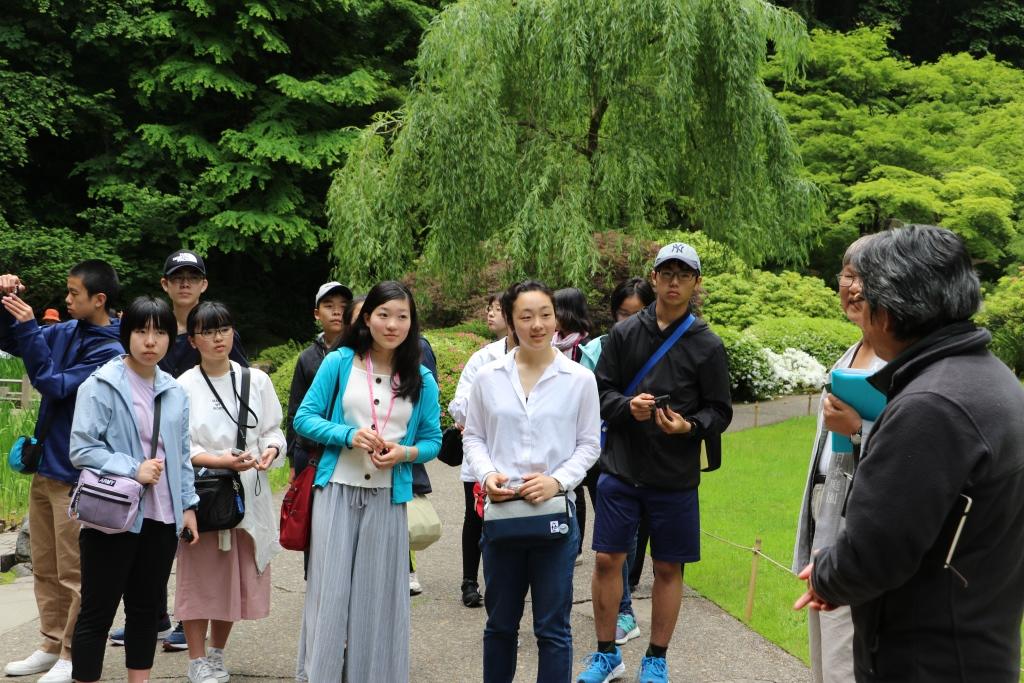 日本庭園にて右側に立つ後ろ姿の男性から説明を受ける生徒たちの写真
