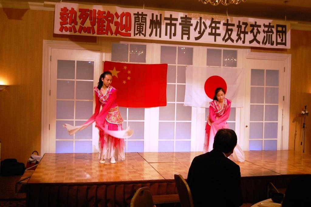 熱烈歓迎蘭州市青少年友好交流団と書かれた舞台上でピンクの衣装を着た女性2名が踊っている写真
