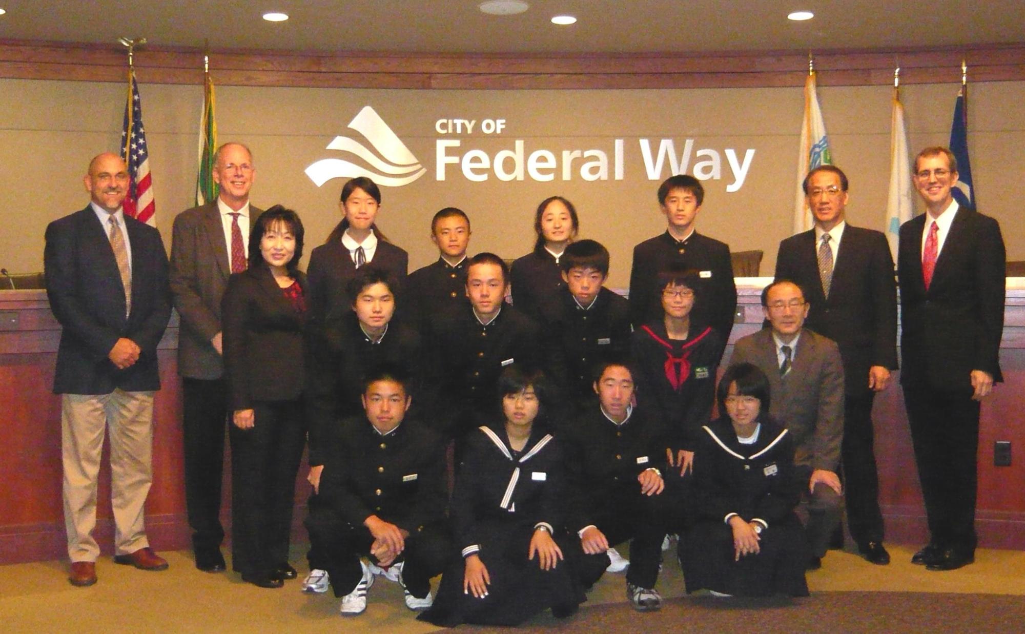 Federalwayと書かれた壁の前で中央に制服を着た生徒たち、両端に外国人たちが立って記念撮影をしている写真