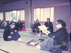 梅香会雅会にて畳の上で、右側で書類を持ち読み上げる男性と、左側で頭を下げる男性の写真