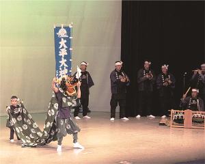 左に獅子舞を披露しており尾の部分に布を持つ子供、右には和楽器を演奏している人が6名写っている写真