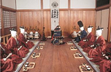 主殿の広間にて赤い衣装をまとい、床に置かれた食事を挟んで平行に並び向かい合って正座している人形7体の写真