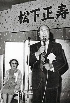 著書『野に叫ぶ声』出版記念会にてマイクを持つ松下 正寿さんと後ろに座る女性の写真