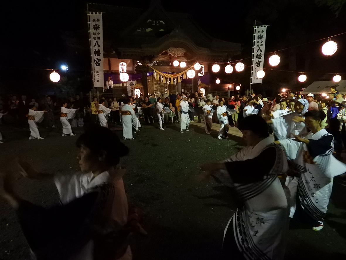 夜の神社の境内にて提灯の明かりの中、円になって盆踊りを踊る人々の写真