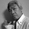 手にお椀と箸を持ち何かを食べようとしカメラ目線をしている田中 稔さんの白黒写真