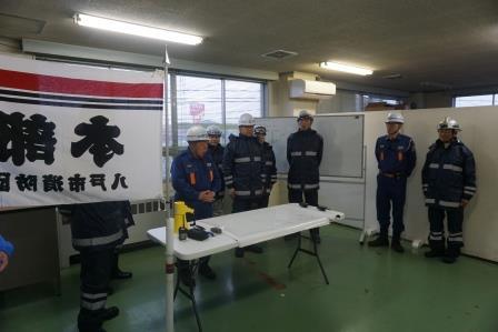 消防署の室内で、テーブルの後ろに立つ上官の消防団員が、訓練の説明を行っている様子の写真