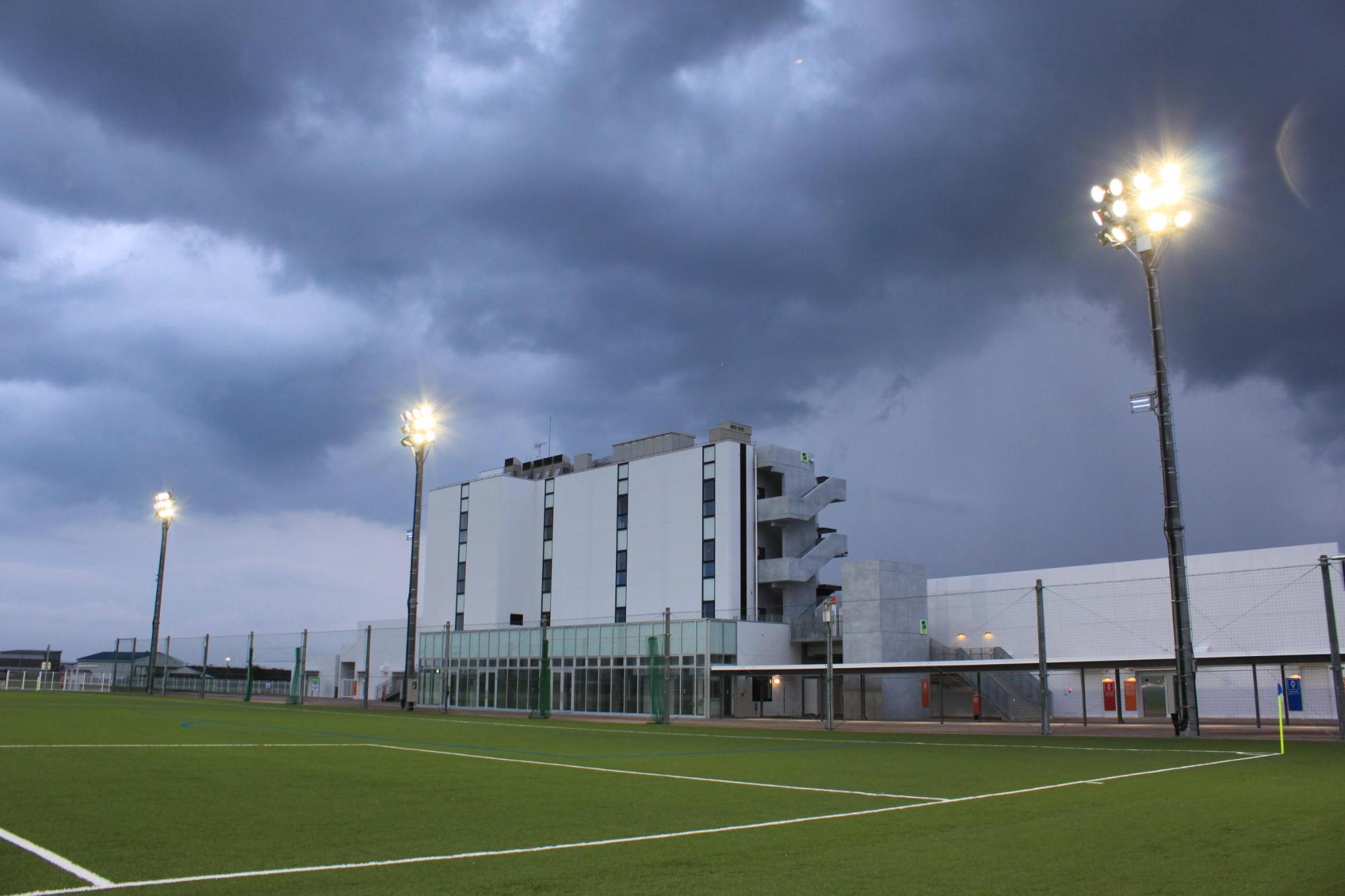 雲が垂れ込め、照明灯が明るく点灯している人工芝球技場のピッチの低い角度からの写真