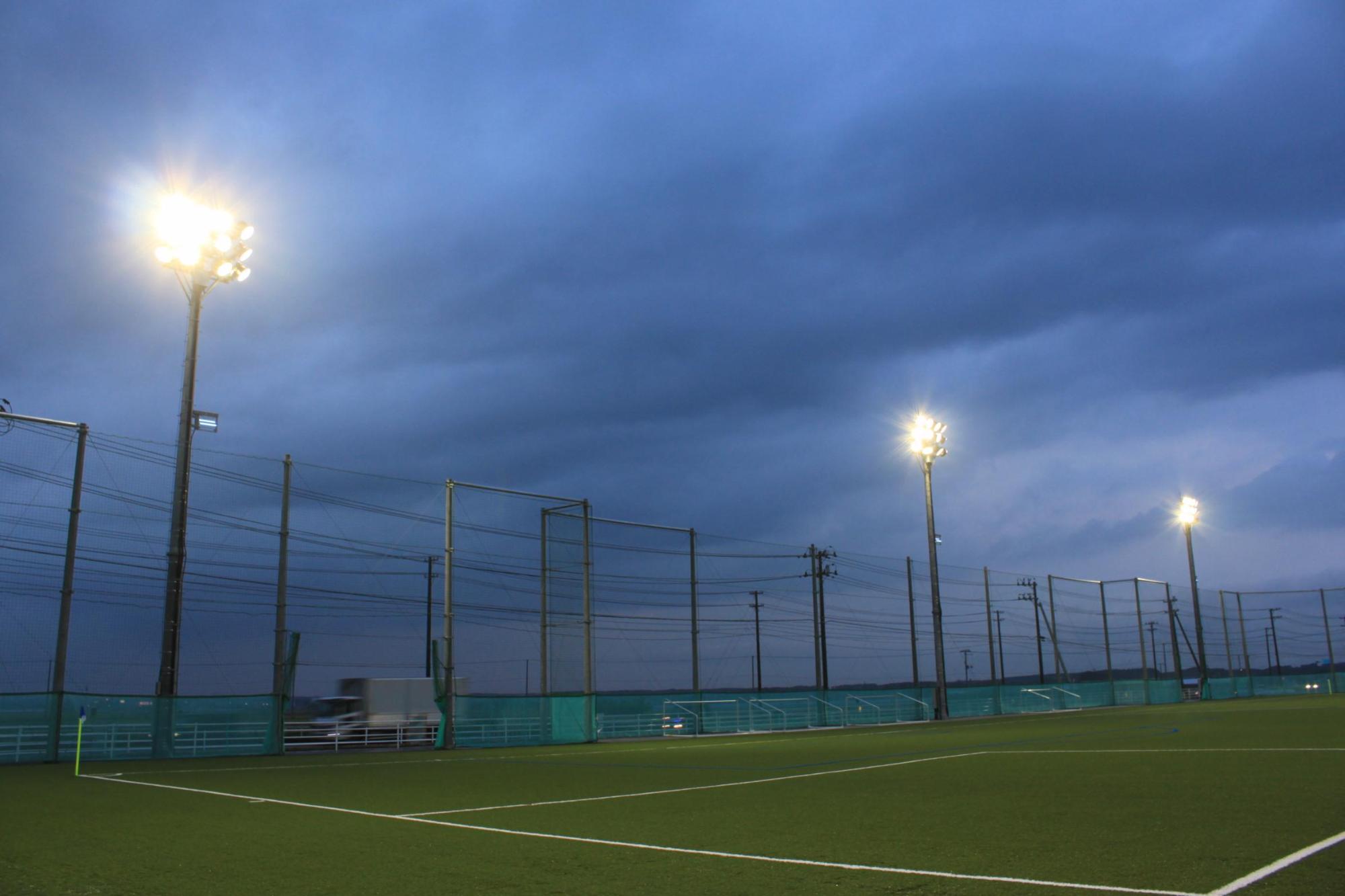 雲が垂れ込め、照明灯が明るく点灯している人工芝球技場のピッチの低い角度からの写真