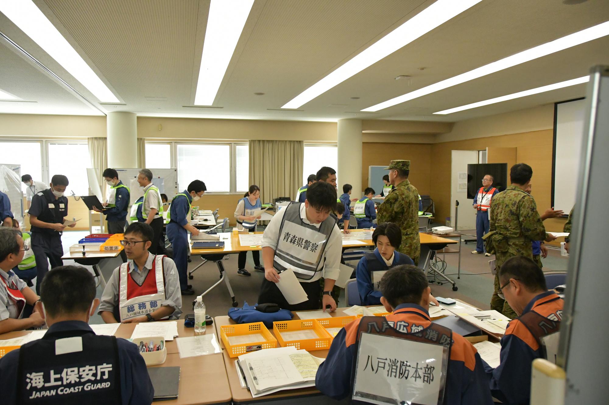会議室で、手前では設置された関係機関用のテーブルでビブスを着た自衛隊、海上保安部、八戸警察署、水道企業団の連絡員がそれぞれ作業をしており、中央では調整会議用のテーブルが設置され、その周辺でビブスを着た数名が作業をしている写真