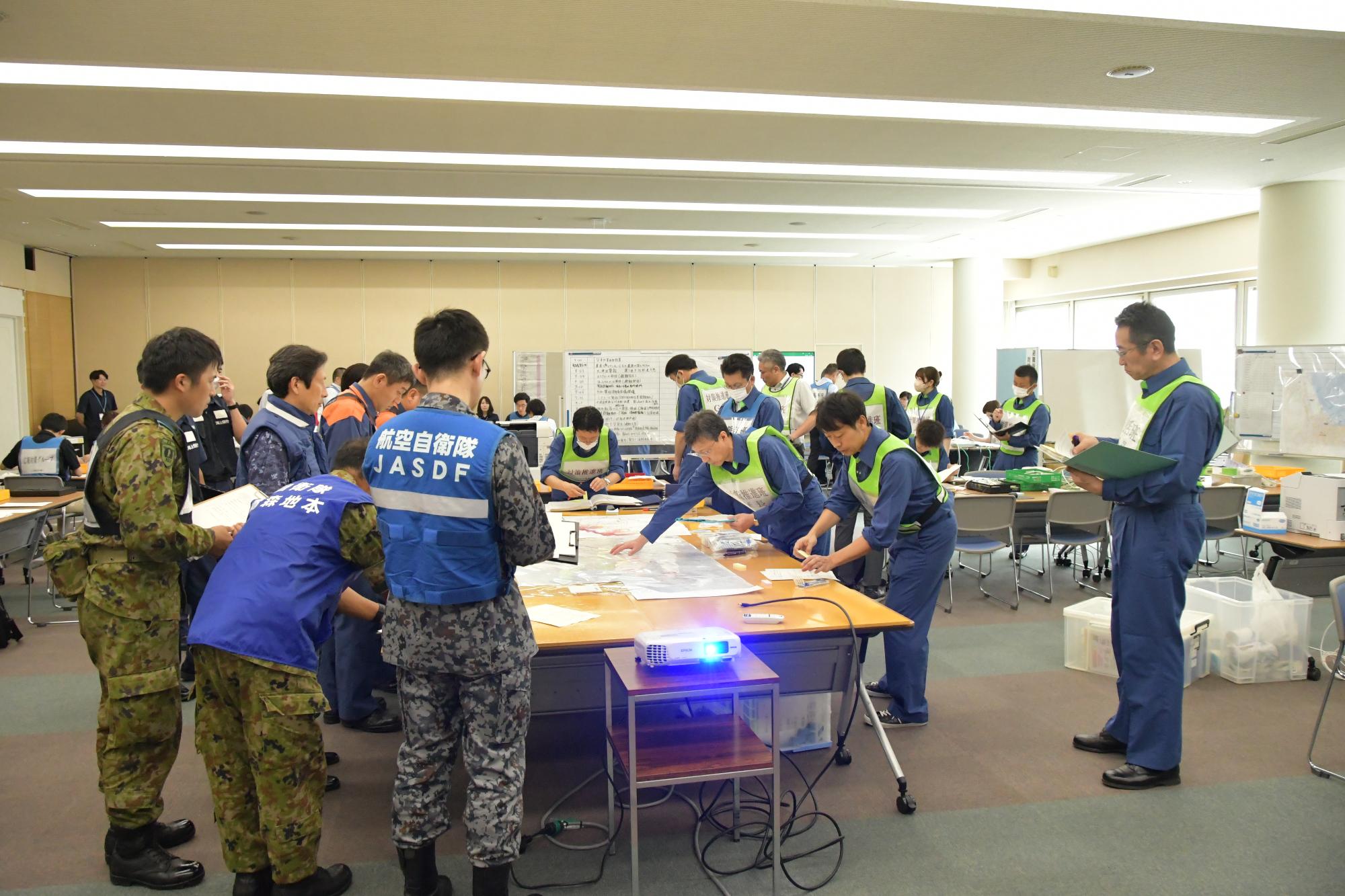 会議室で、中央に設置されたテーブルにビブスを着た市の職員と自衛隊、消防本部などの関係機関が集まり、テーブルに置かれた地図を囲み、話し合っている写真