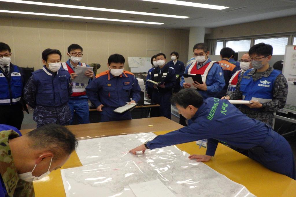 会議室の中央に地図を広げたテーブルで、自衛隊、消防、警察などの担当者が地図を囲んで集まっており、市の職員が左手で地図を示し、説明している写真