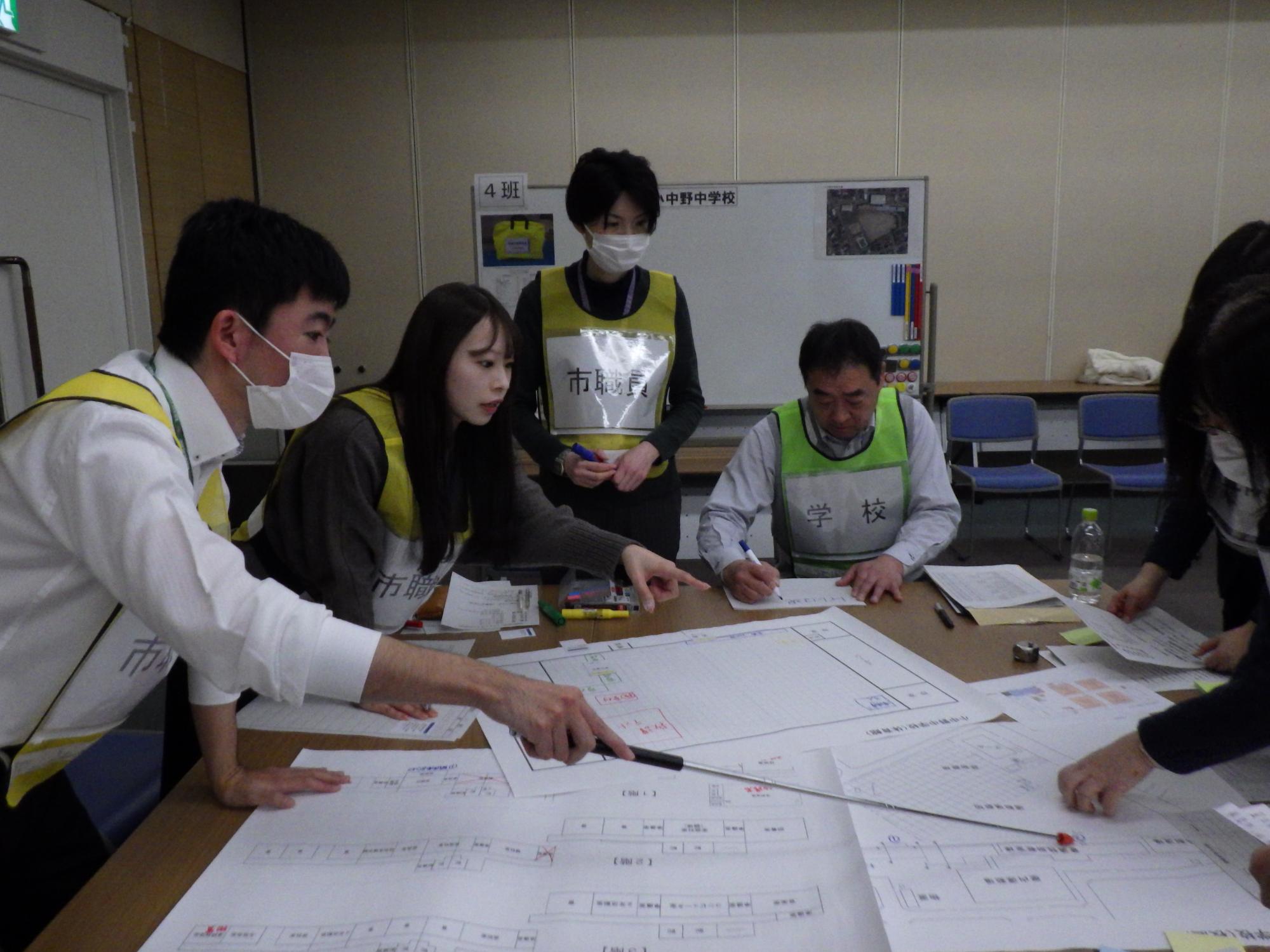 会議室でビブスを着けた参加者6名が、テーブルに広げた学校の平面図を囲み、話し合っており、そのうち1名が指示棒を使って平面図の一部を示し、隣の参加者も同じ方向を指で示している写真