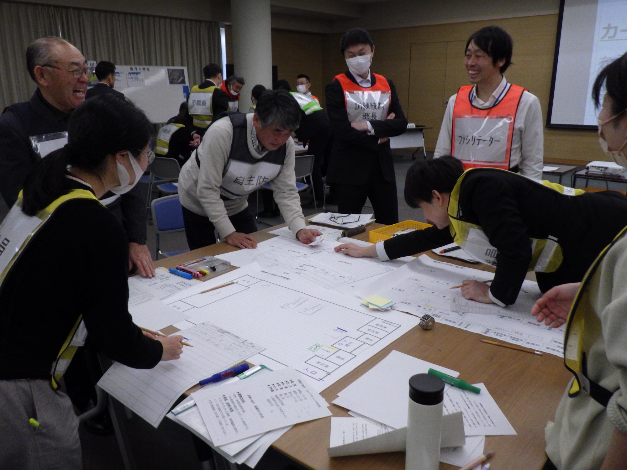 会議室でビブスを着けた参加者6名が、テーブルに広げた学校の平面図を囲み、うち2名が平面図に訓練カードをのせ、その他の者は立った姿勢で話し合っている写真